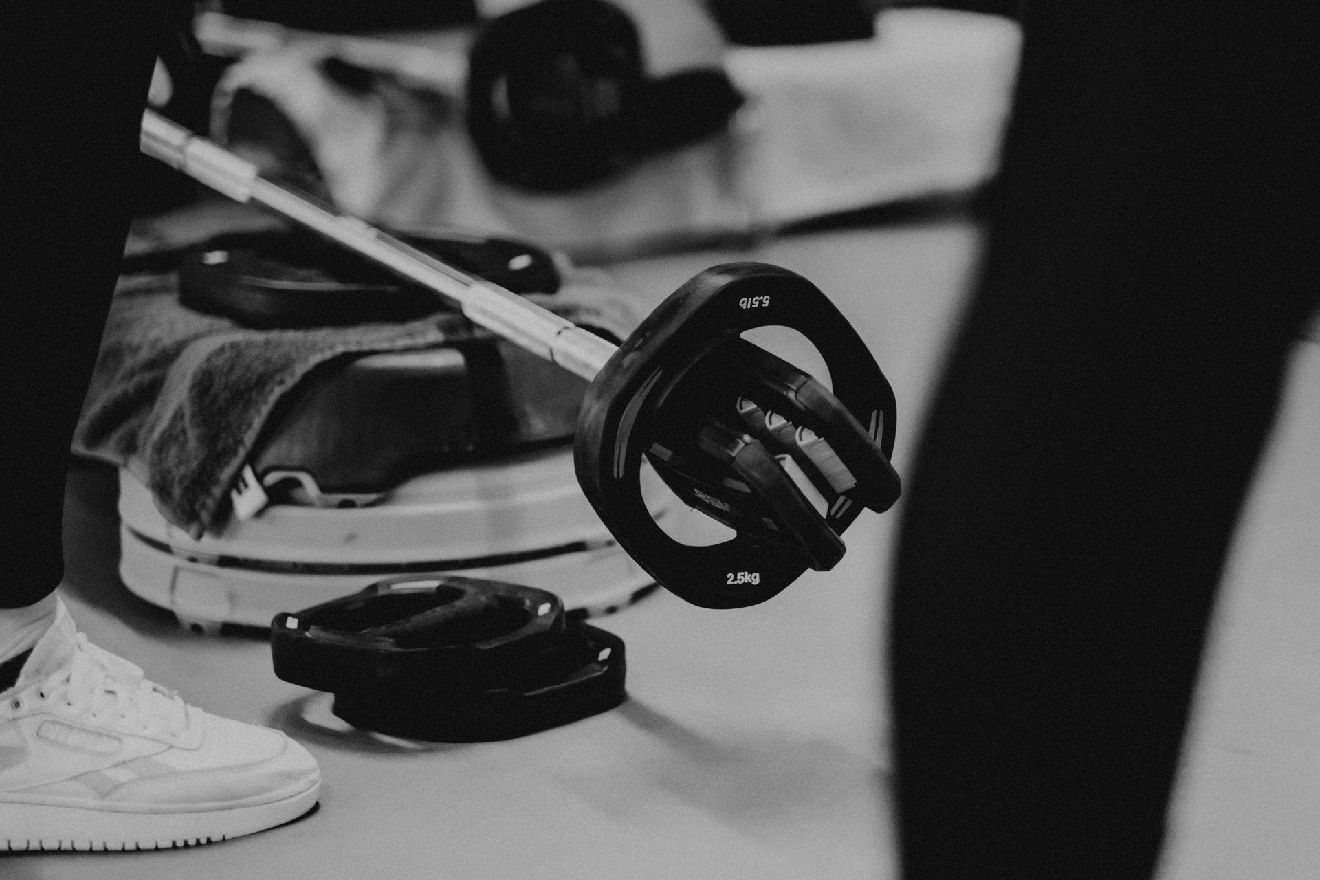 Body Pump en pump your bum materialen liggen op de grond: een step, handoek, gewichten en een barbell. Sporter staat er naast. We zien diens voeten.